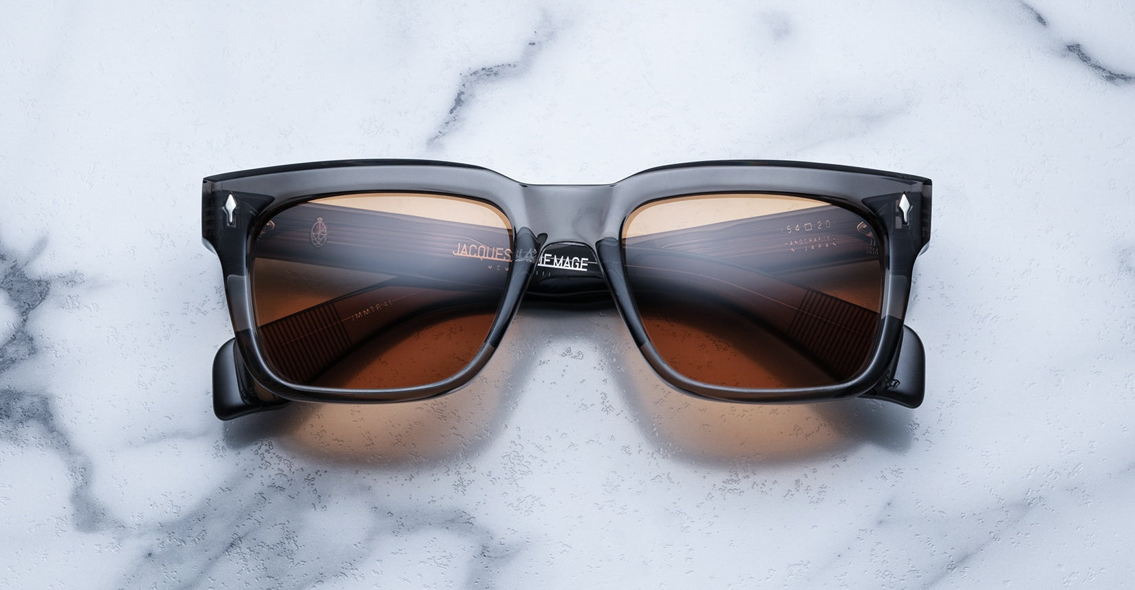 The Jacques Marie Mage Torino eyewear features bold rectangular acetate frames with brown-tinted lenses, showcased on a white marble surface.