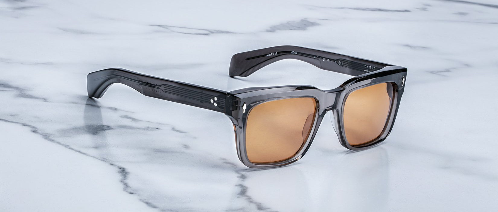 Jacques Marie Mage Torino sunglasses by Jacques Marie Mage feature bold, square dark gray acetate frames and brown-tinted lenses, showcased on a white marble surface—a striking example of handcrafted eyewear.