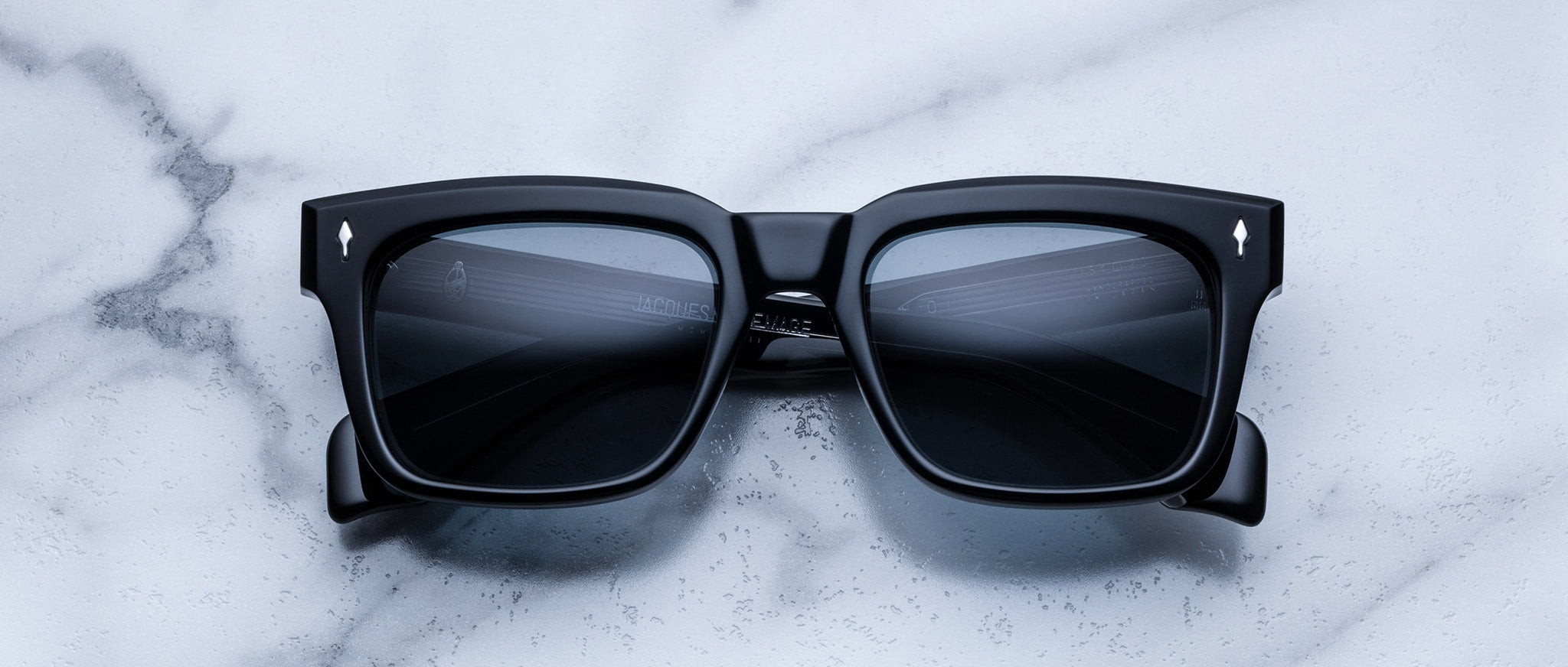 Jacques Marie Mage Torino sunglasses by Jacques Marie Mage feature bold black square frames with dark lenses, inspired by classic rectangular eyeglass styles, showcased on a white marble surface—modern and reminiscent of handcrafted acetate design.
