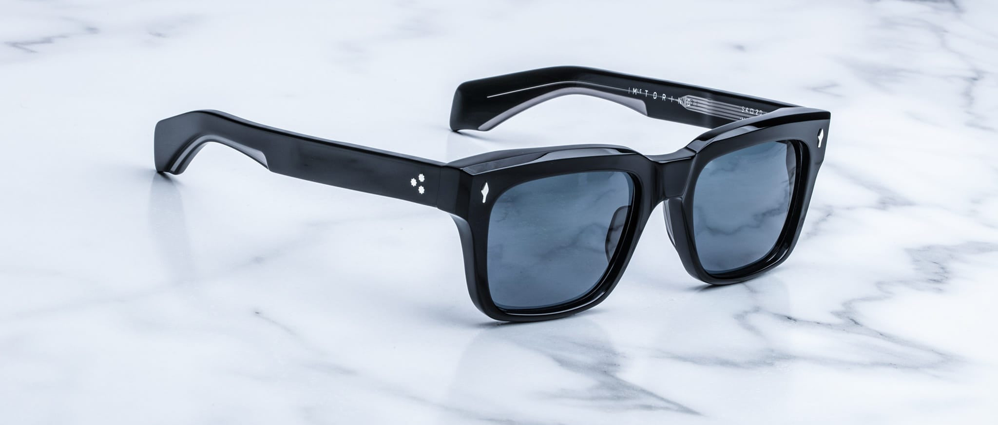 Jacques Marie Mage Torino sunglasses feature bold, black handcrafted acetate frames and dark lenses, resting on a white marble surface. Inspired by iconic style, these Jacques Marie Mage rectangular sunglasses exude timeless elegance.