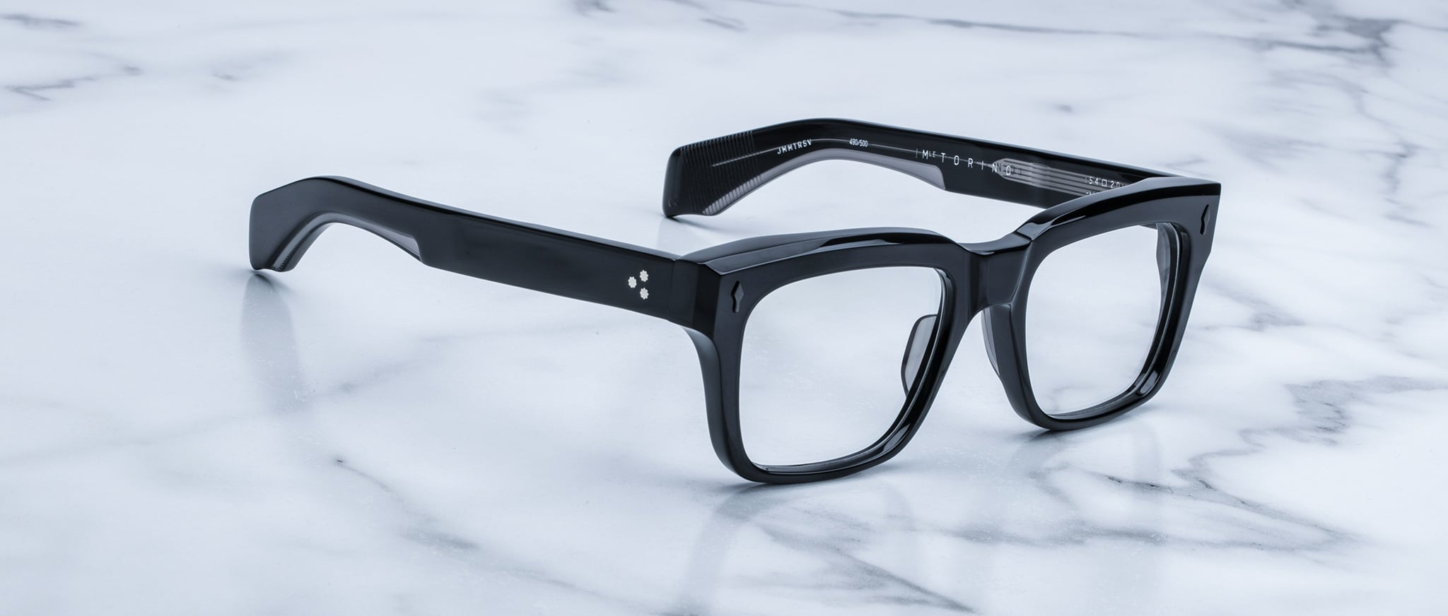 A pair of Jacques Marie Mage Torino eyeglass frames by Jacques Marie Mage sit on a white marble surface, highlighting the brand's handcrafted acetate and bold rectangular design.