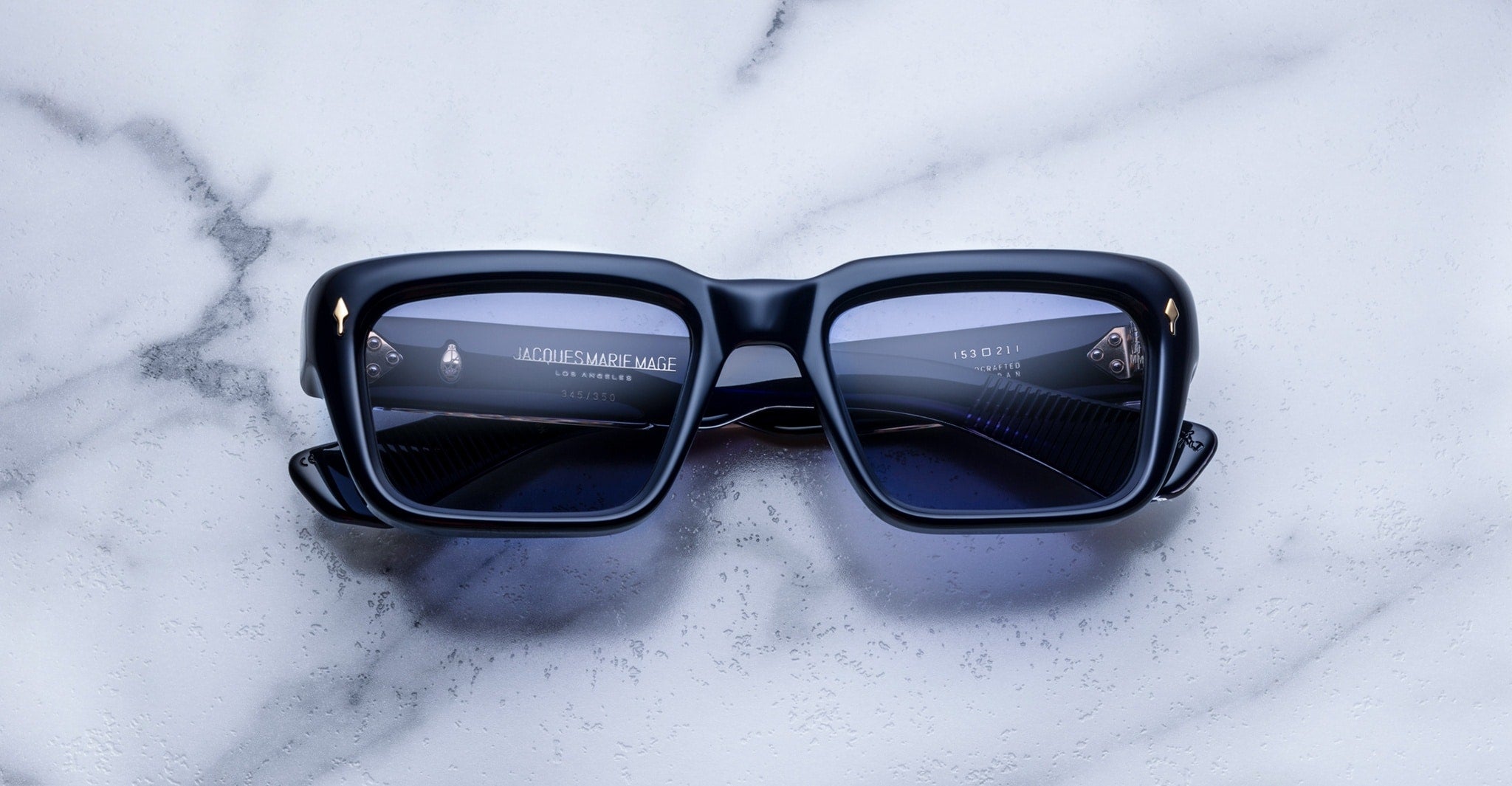 Black rectangular sunglasses with blue-tinted lenses rest on a white marble surface. The lenses reflect a modern interior scene with text and geometric shapes visible in the reflection.