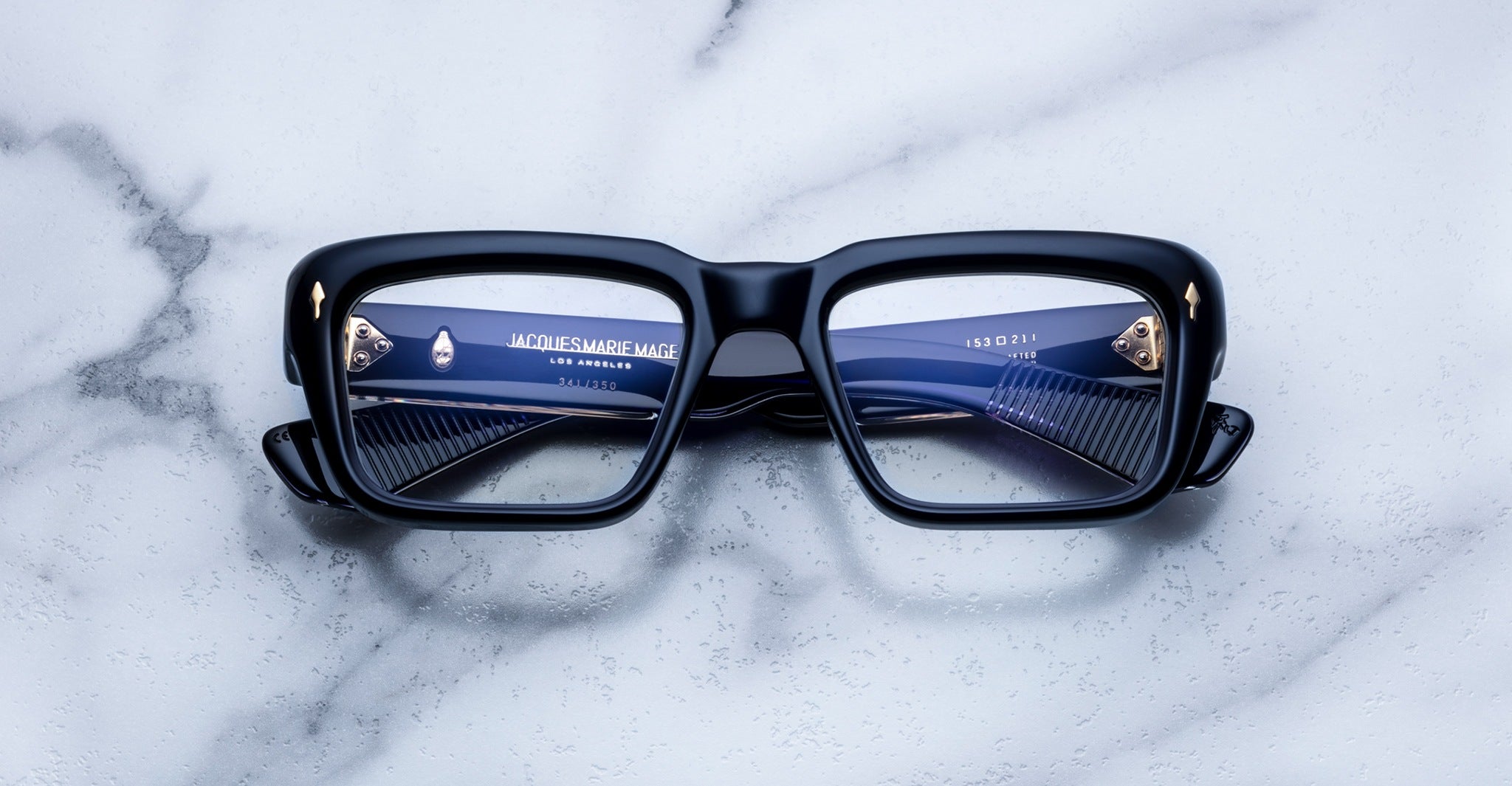 Thick black rectangular eyeglasses are placed face up on a white marble surface, with blue details and reflections visible on the lenses and inside the frame arms.