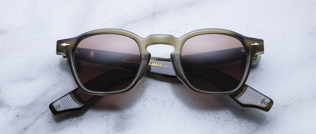 A pair of Jacques Marie Mage Zephirin sunglasses, featuring square frames with dark lenses and translucent grayish handcrafted acetate rims, sits on a white marble surface, highlighting the brand’s limited edition sophistication.
