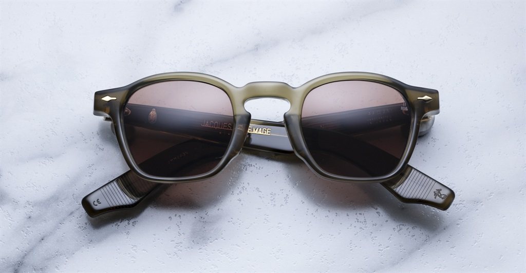 A pair of Jacques Marie Mage Zephirin sunglasses, featuring square frames with dark lenses and translucent grayish handcrafted acetate rims, sits on a white marble surface, highlighting the brand’s limited edition sophistication.