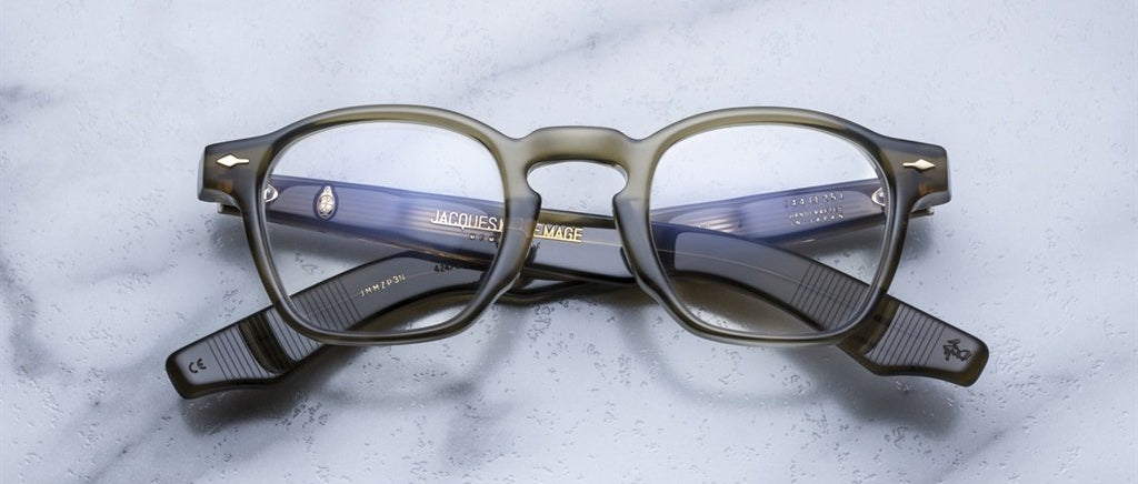 A pair of Jacques Marie Mage Zephirin round dark-framed acetate eyeglasses with blue-tinted arms by Jacques Marie Mage, displayed on a light marble surface.