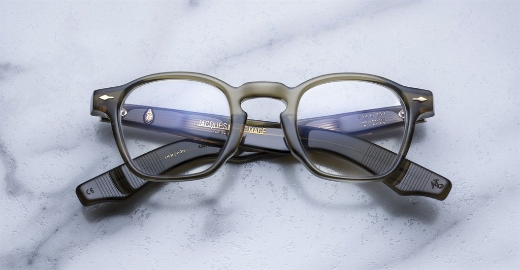 A pair of Jacques Marie Mage Zephirin round dark-framed acetate eyeglasses with blue-tinted arms by Jacques Marie Mage, displayed on a light marble surface.
