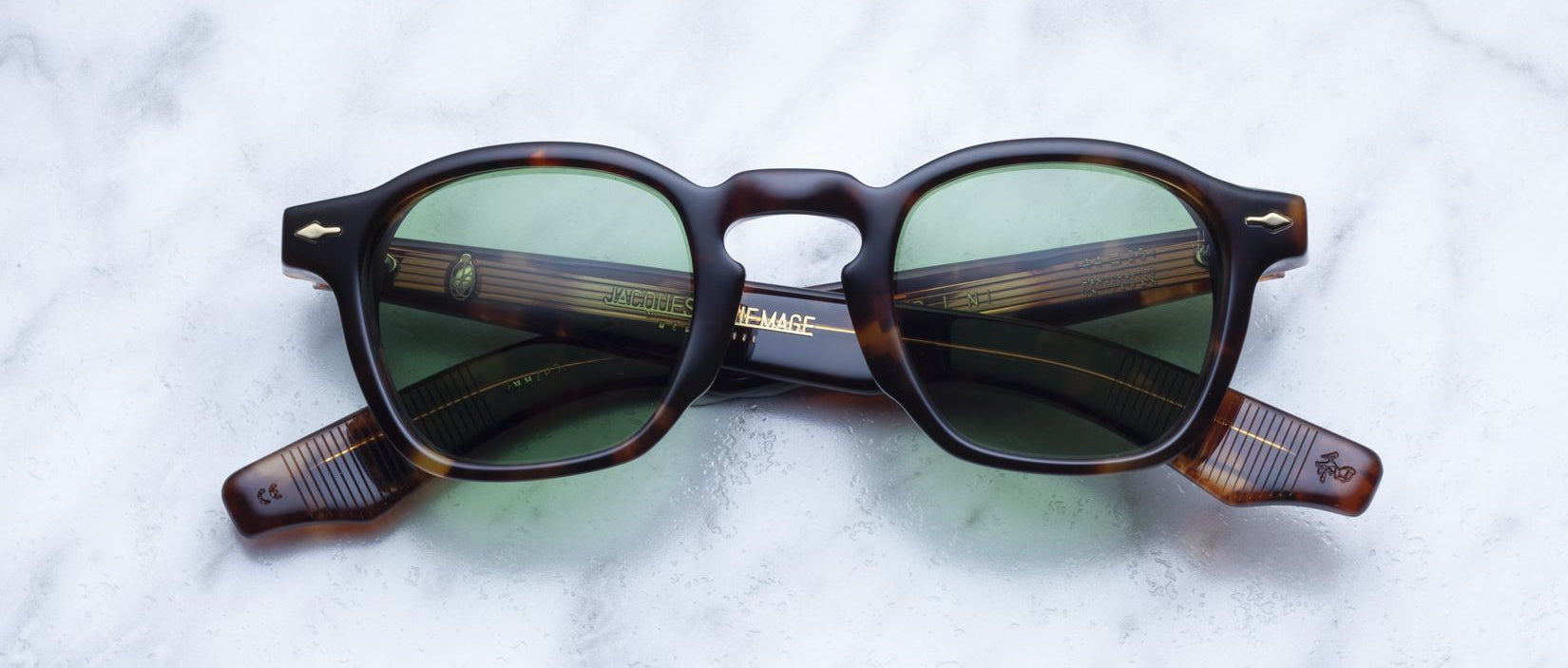 Jacques Marie Mage Zephirin square sunglasses in tortoiseshell acetate with green lenses are elegantly displayed on a light marble surface.