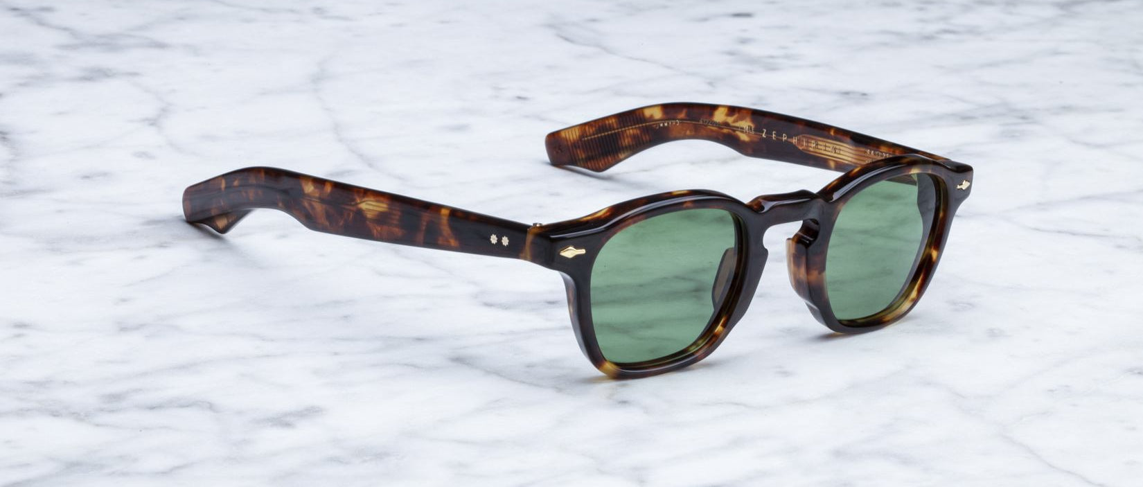 Jacques Marie Mage Zephirin tortoiseshell sunglasses with green lenses rest on white marble—a limited edition, handcrafted acetate eyewear piece by Jacques Marie Mage.