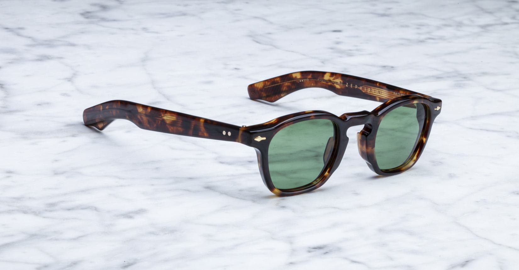 Jacques Marie Mage Zephirin tortoiseshell sunglasses with green lenses rest on white marble—a limited edition, handcrafted acetate eyewear piece by Jacques Marie Mage.