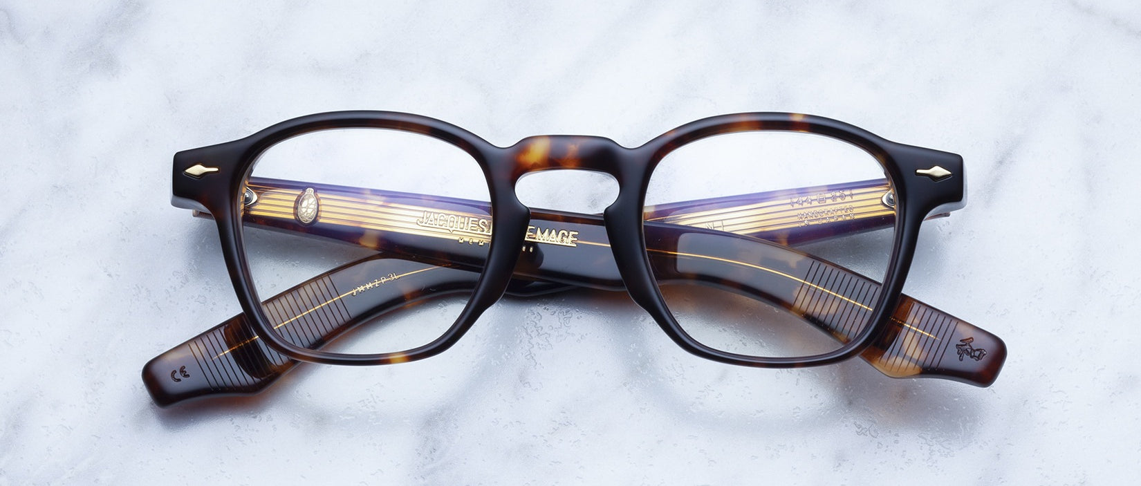 A pair of Jacques Marie Mage Zephirin rectangular eyeglasses with tortoiseshell acetate frames rests on a marble surface, showcasing the brand's handcrafted design.