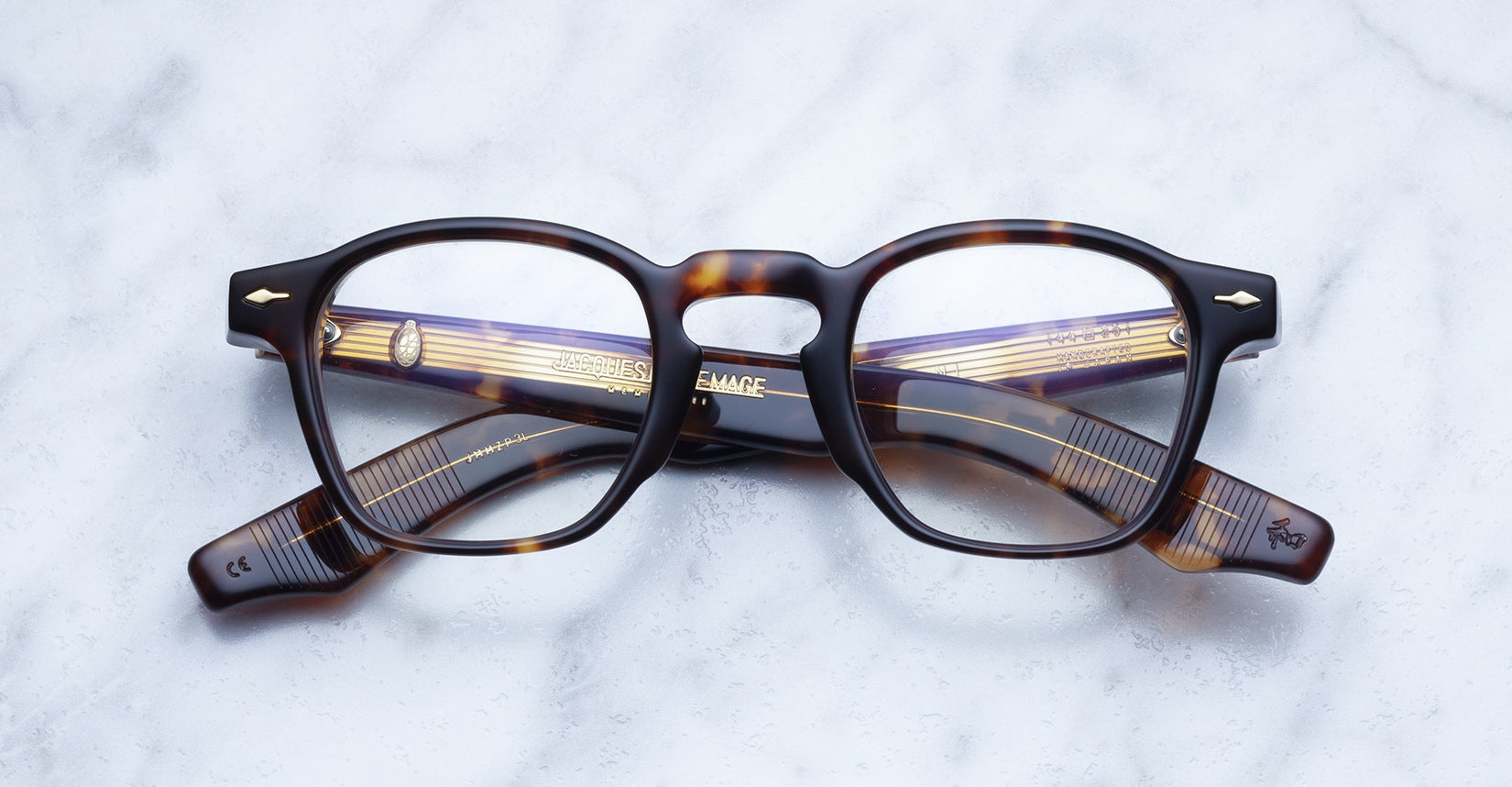 A pair of Jacques Marie Mage Zephirin rectangular eyeglasses with tortoiseshell acetate frames rests on a marble surface, showcasing the brand's handcrafted design.