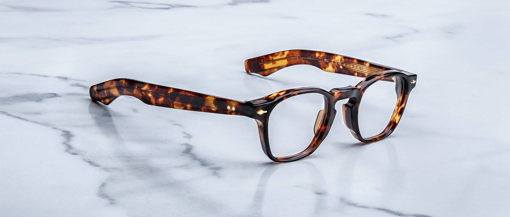 A pair of Jacques Marie Mage Zephirin handcrafted acetate spectacles with round lenses rests on a white marble surface.