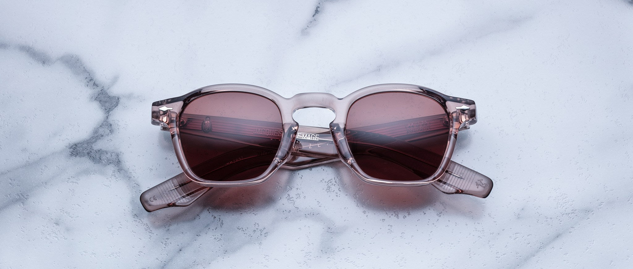 Handcrafted Jacques Marie Mage Zephirin sunglasses, featuring clear acetate frames and pink tinted lenses, rest on white marble—a tribute to the brand’s iconic limited edition eyewear.