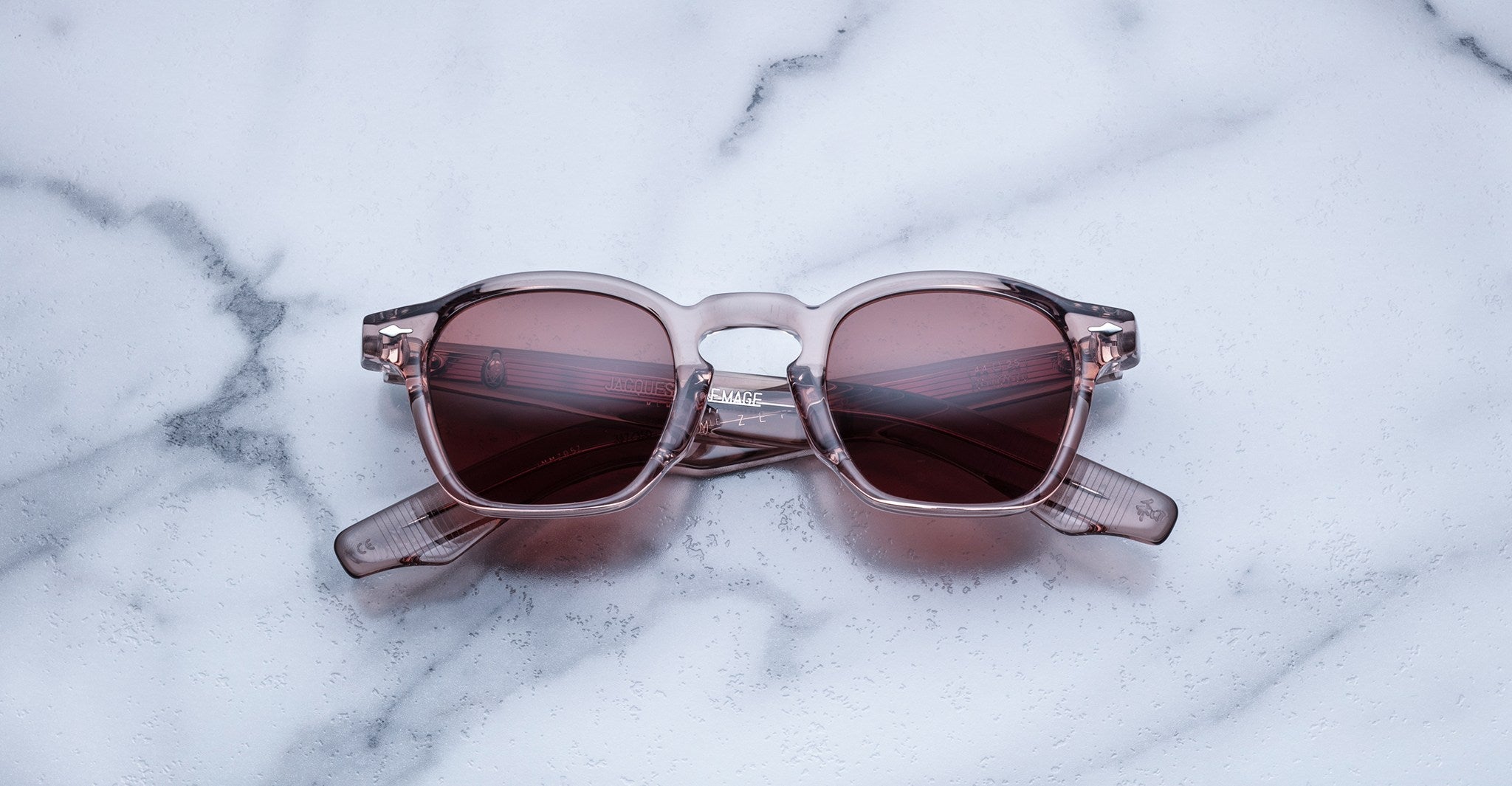 Handcrafted Jacques Marie Mage Zephirin sunglasses, featuring clear acetate frames and pink tinted lenses, rest on white marble—a tribute to the brand’s iconic limited edition eyewear.