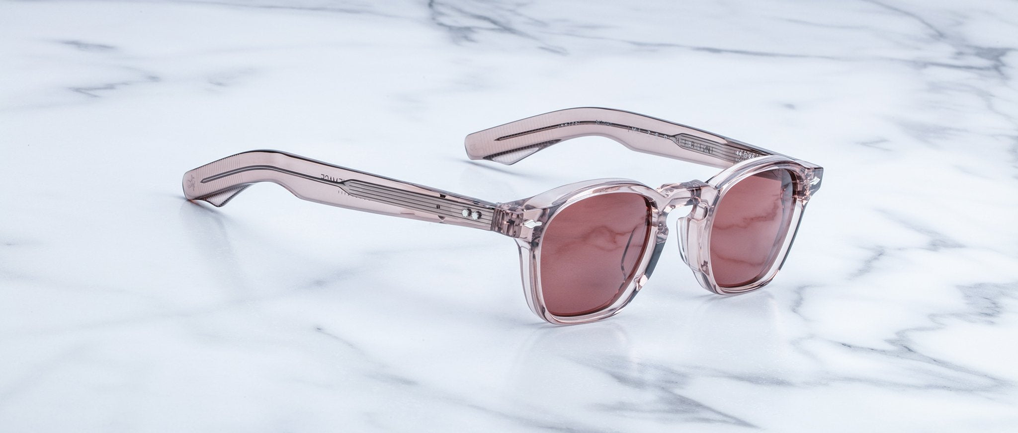 The Jacques Marie Mage Zephirin, limited edition sunglasses with transparent pink acetate frames, rest on a white marble surface, highlighting the brand’s handcrafted elegance.