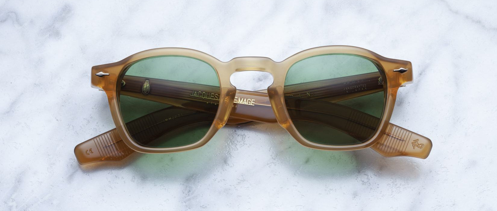 Jacques Marie Mage Zephirin sunglasses in brown translucent acetate with green lenses, a limited edition handcrafted by Jacques Marie Mage, are displayed folded on a white marble surface—an exquisite eyewear piece.