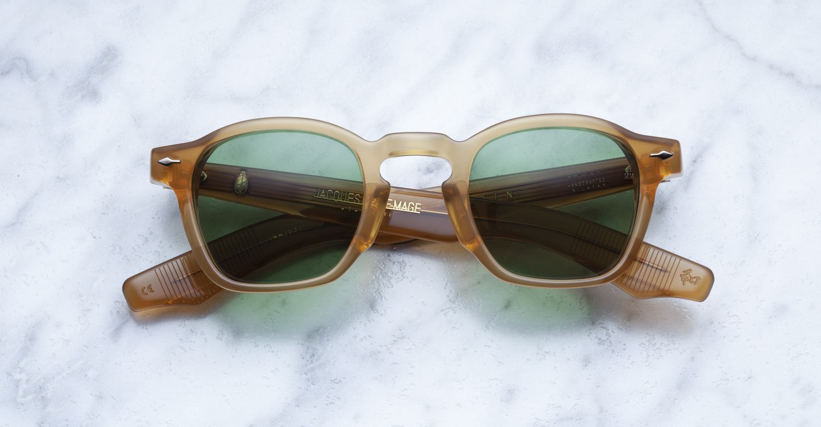 Jacques Marie Mage Zephirin sunglasses in brown translucent acetate with green lenses, a limited edition handcrafted by Jacques Marie Mage, are displayed folded on a white marble surface—an exquisite eyewear piece.