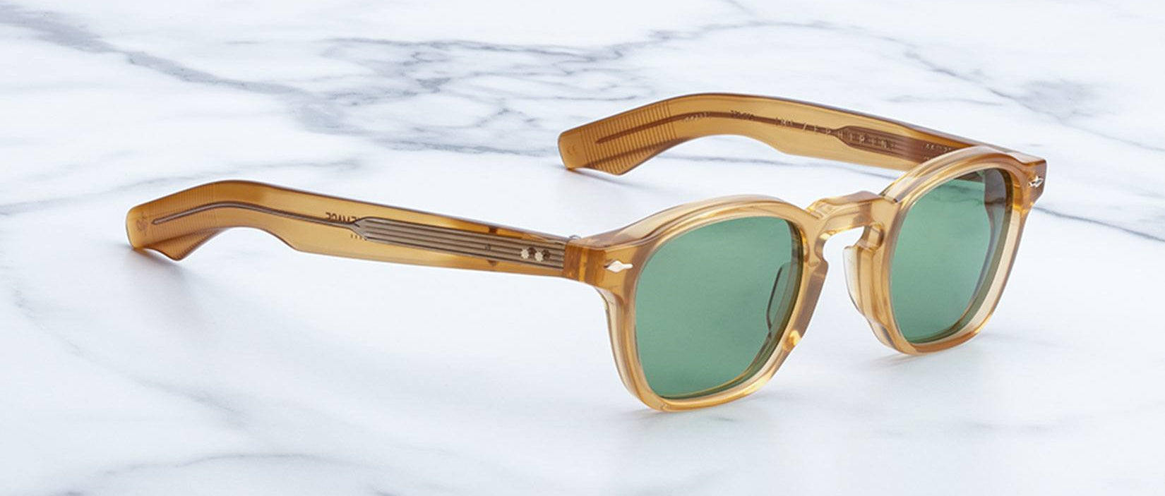 A pair of Jacques Marie Mage Zephirin sunglasses featuring amber square acetate frames and green lenses, displayed on a white marble surface.