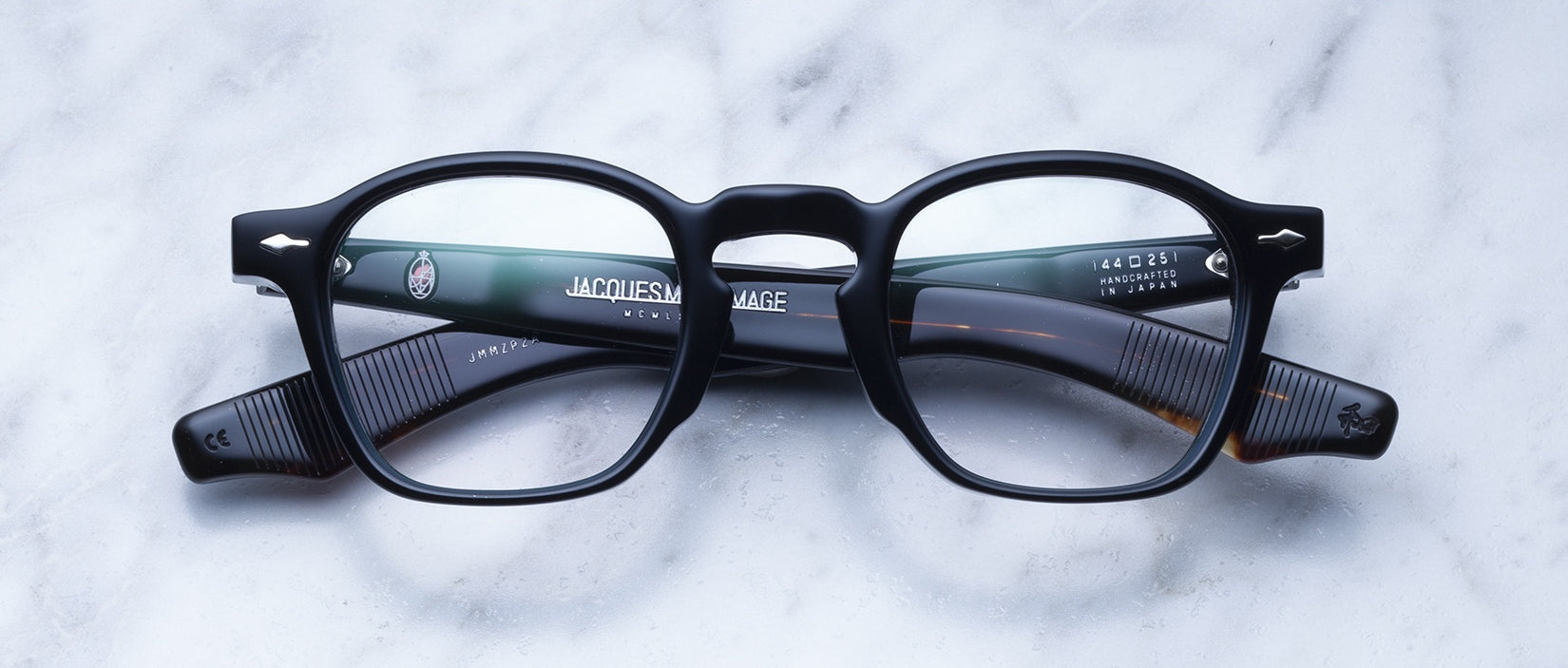A pair of Jacques Marie Mage Zephirin black round eyeglasses with thick handcrafted acetate frames and ribbed temple arms rests on a white marble surface.