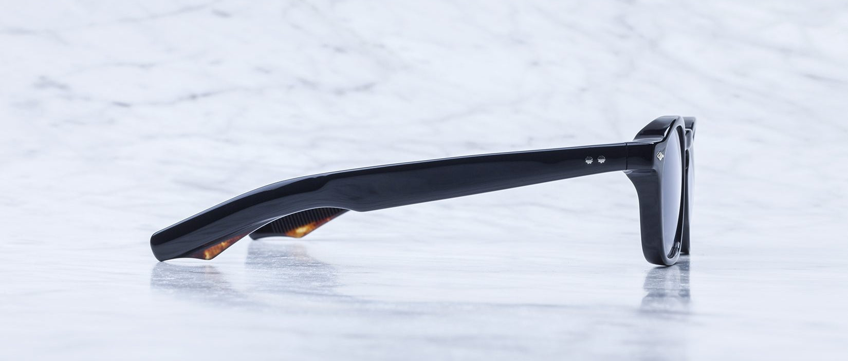 A pair of Jacques Marie Mage Zephirin sunglasses, shown from the side with dark lenses, rest on a light marble surface—handcrafted Jacques Marie Mage acetate eyewear for discerning tastes.