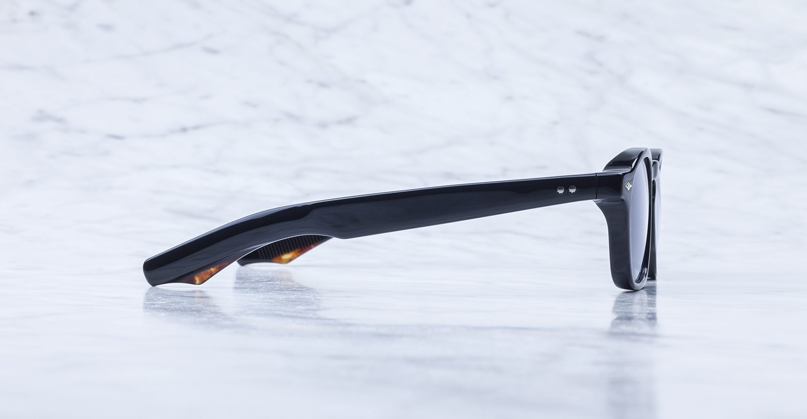 A pair of Jacques Marie Mage Zephirin sunglasses, shown from the side with dark lenses, rest on a light marble surface—handcrafted Jacques Marie Mage acetate eyewear for discerning tastes.