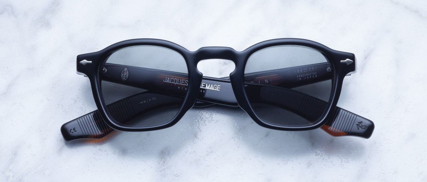 A pair of Jacques Marie Mage Zephirin black acetate sunglasses with dark lenses from Jacques Marie Mage is displayed on a white marble surface.