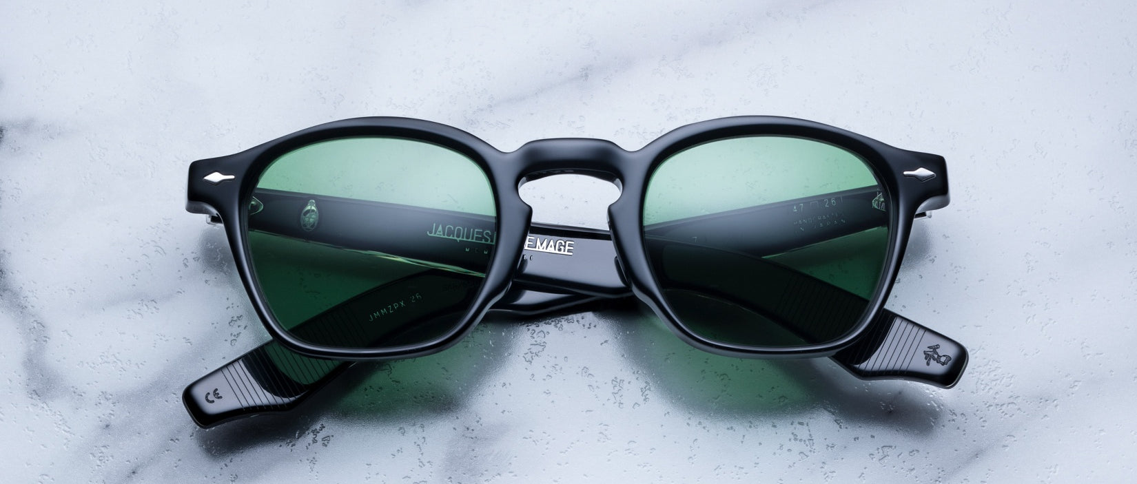 A pair of Jacques Marie Mage Zephirin round sunglasses with green-tinted lenses is displayed on a white marble surface, highlighting the exclusivity of this limited edition eyewear by Jacques Marie Mage.