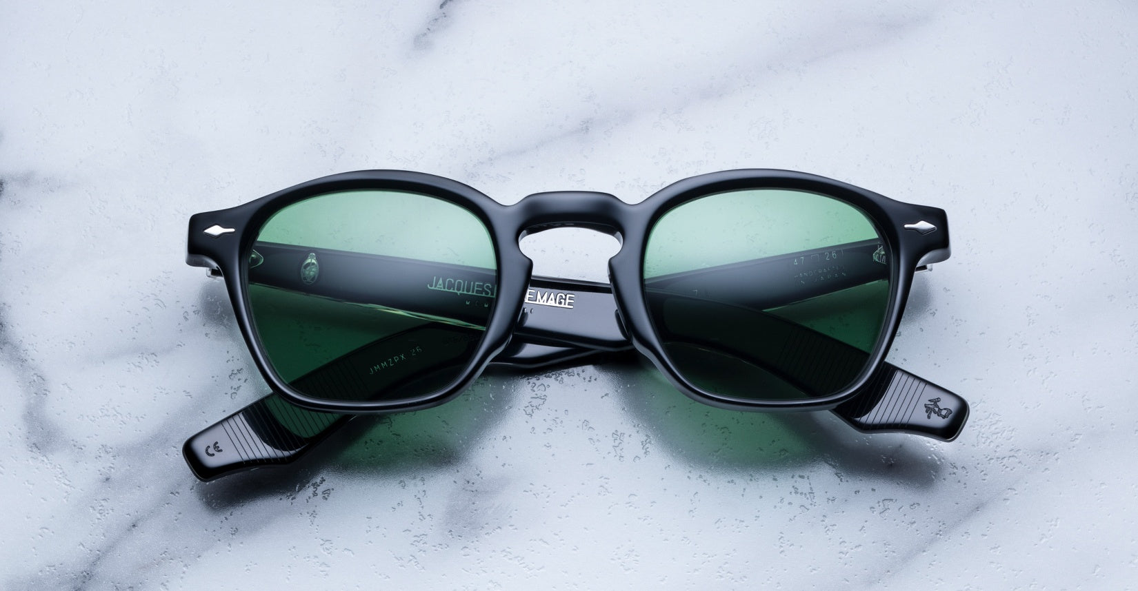 A pair of Jacques Marie Mage Zephirin round sunglasses with green-tinted lenses is displayed on a white marble surface, highlighting the exclusivity of this limited edition eyewear by Jacques Marie Mage.