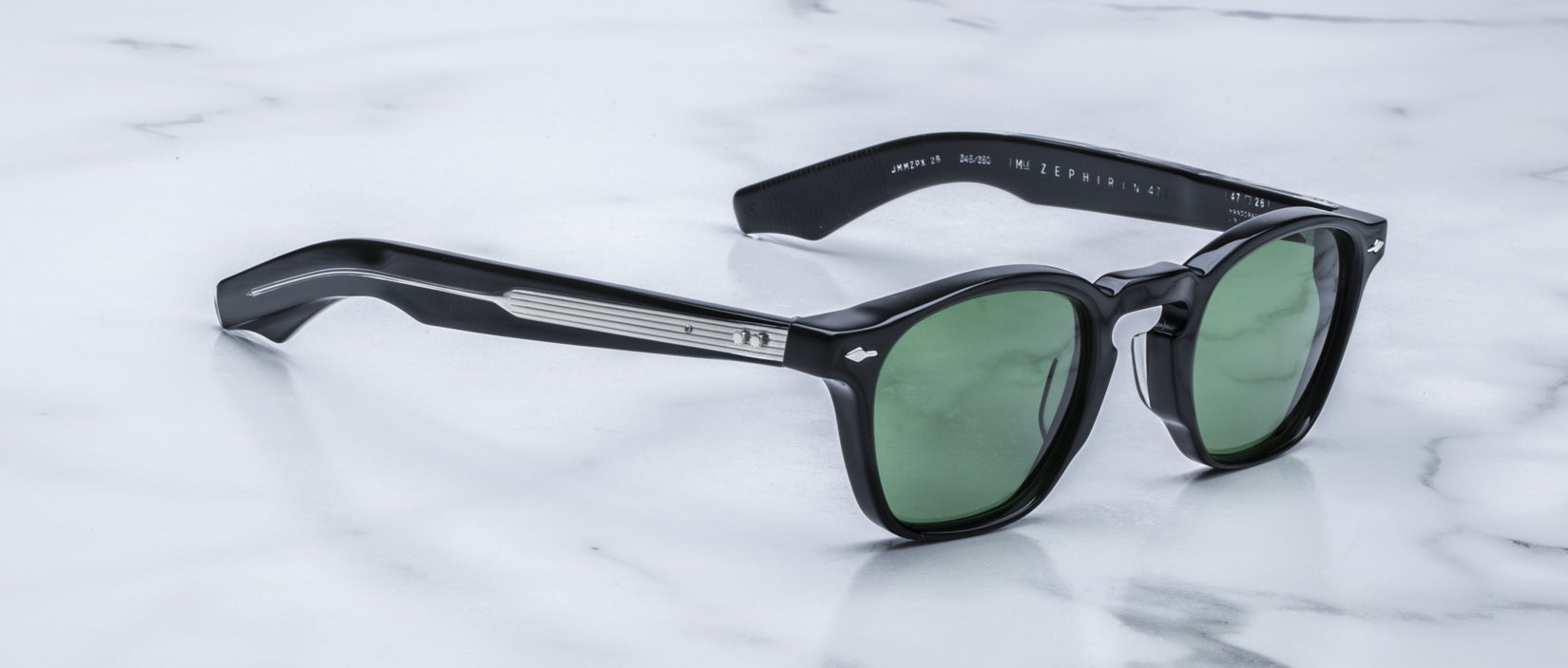 Jacques Marie Mage Zephirin sunglasses with black frames and green lenses rest on white marble, highlighting the exclusive appeal of this limited edition eyewear by Jacques Marie Mage.