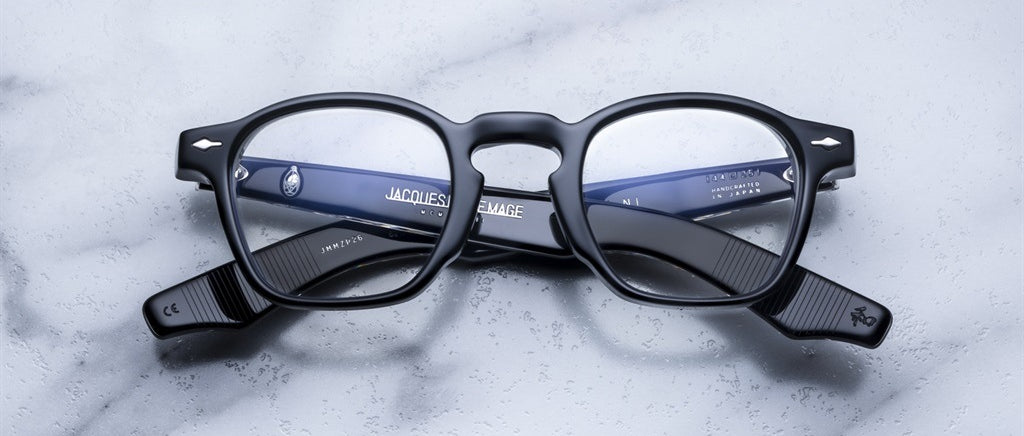 A pair of Jacques Marie Mage Zephirin spectacles with bold black acetate frames rests on marble, their temples crossed—showcasing the brand’s limited edition craftsmanship.