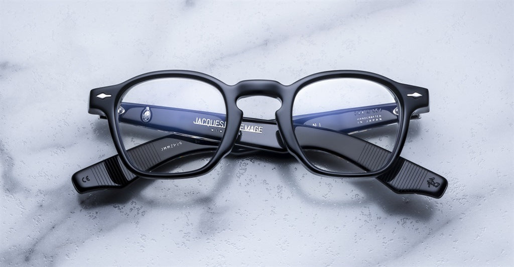 A pair of Jacques Marie Mage Zephirin spectacles with bold black acetate frames rests on marble, their temples crossed—showcasing the brand’s limited edition craftsmanship.