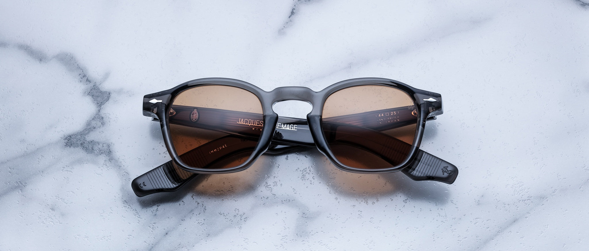 A pair of Jacques Marie Mage Zephirin sunglasses with brown lenses and a dark frame sits on white marble, highlighting the exclusive appeal of this limited edition by Jacques Marie Mage.