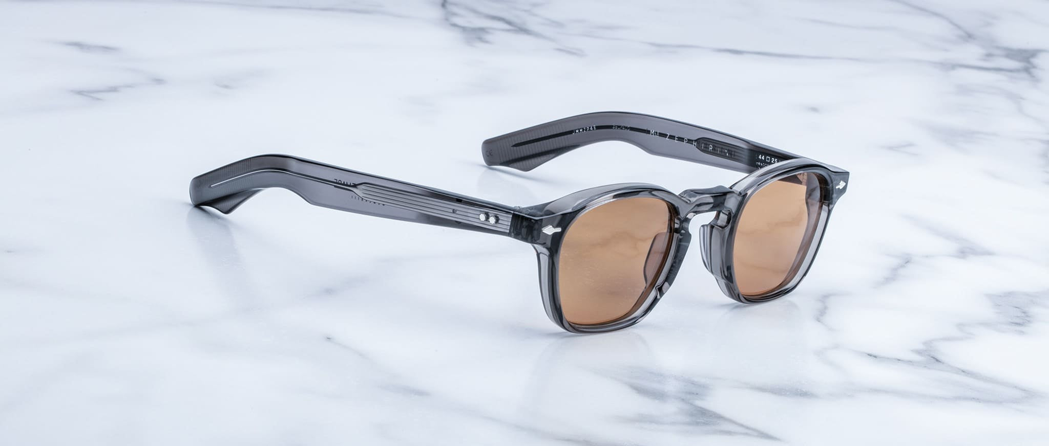 The Jacques Marie Mage Zephirin sunglasses, featuring brown lenses and thick translucent gray frames, are displayed elegantly on a white marble surface.
