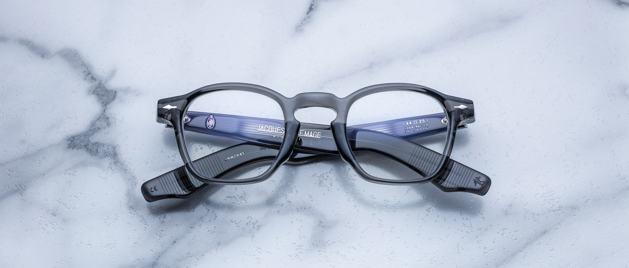 A pair of Jacques Marie Mage Zephirin eyeglasses with black acetate frames is displayed upside down on a white marble surface.