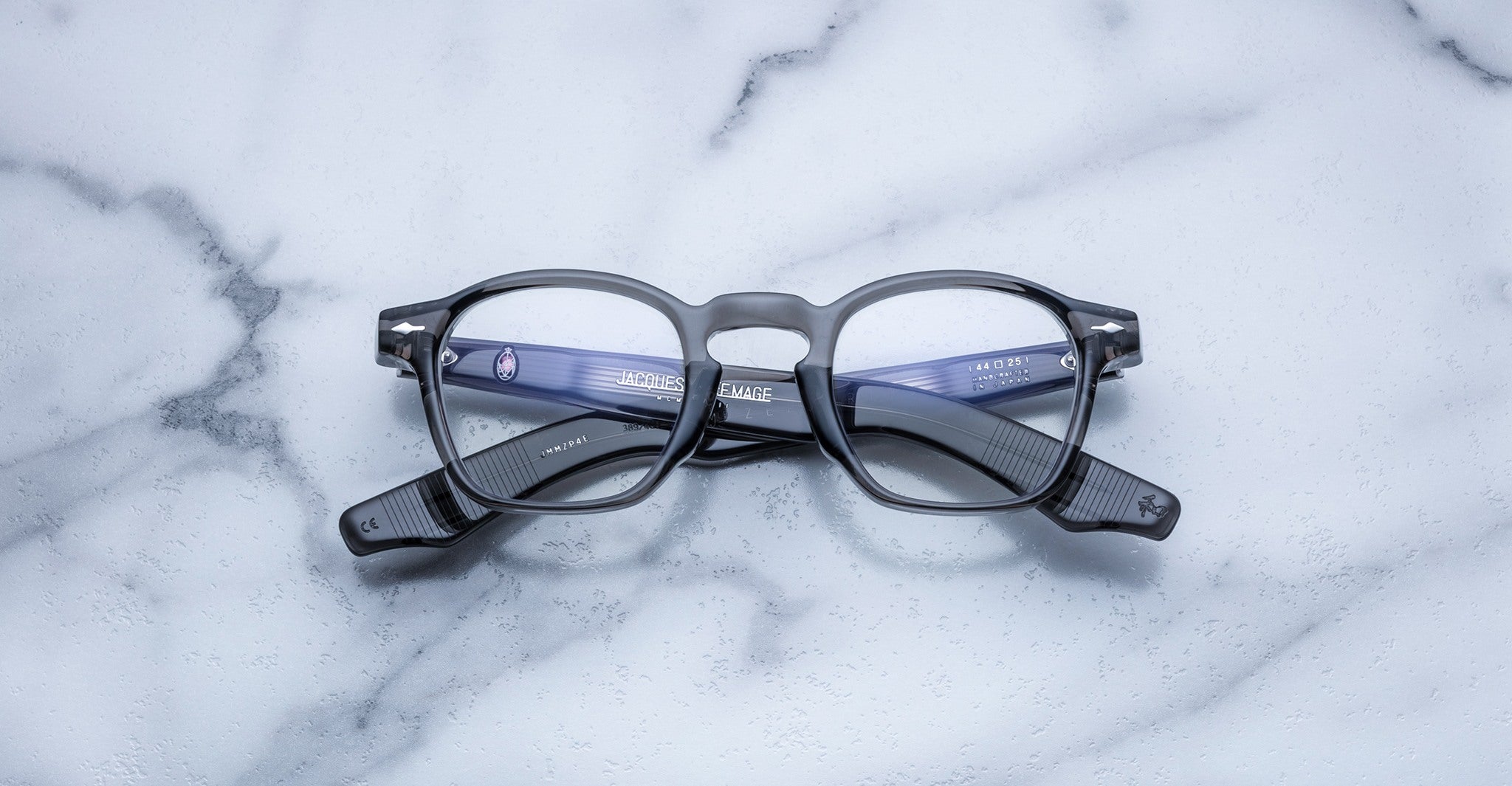 A pair of Jacques Marie Mage Zephirin eyeglasses with black acetate frames is displayed upside down on a white marble surface.