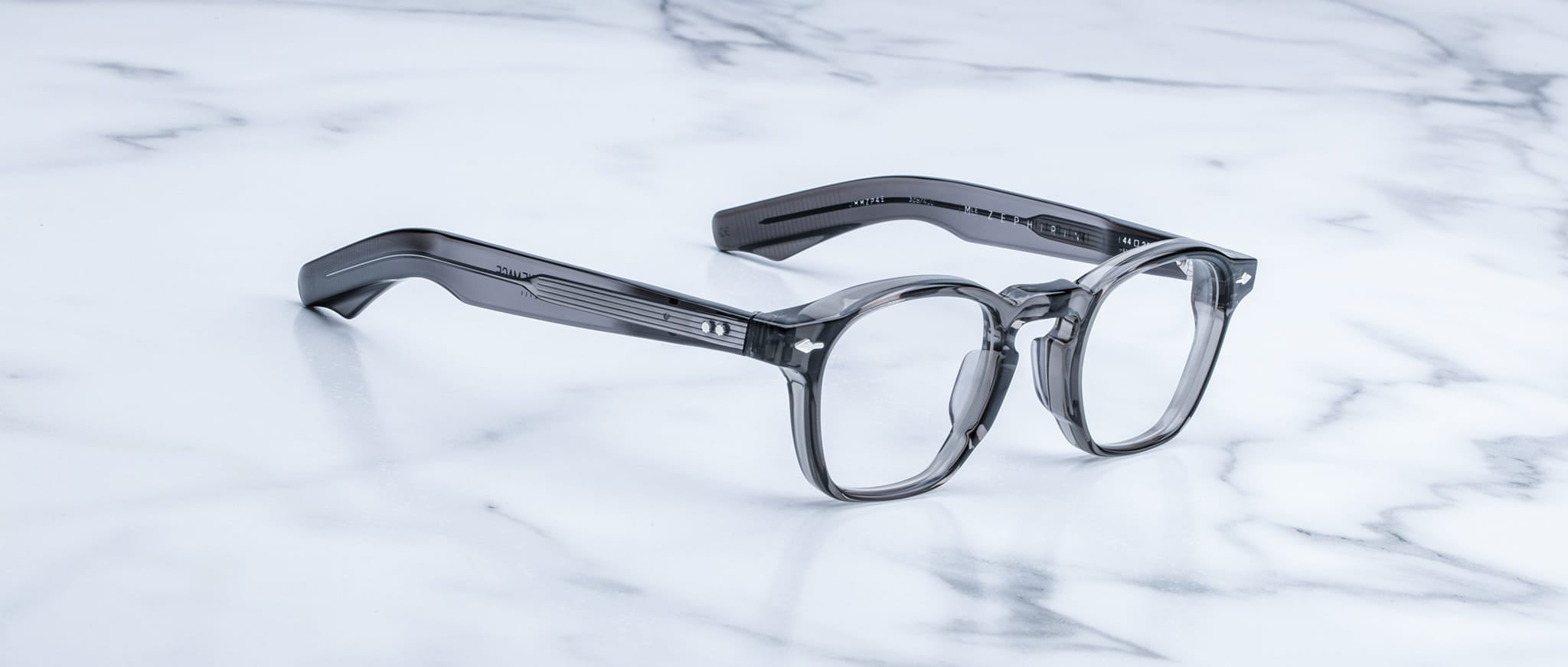 The Jacques Marie Mage Zephirin, transparent gray handcrafted acetate spectacles with thick frames, rest on a white marble surface.