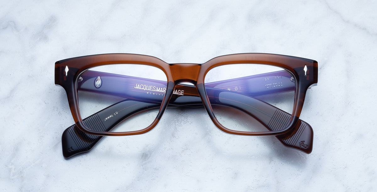 The Jacques Marie Mage Molino eyeglasses in Hickory feature thick, rectangular brown and translucent purple frames. Resting on a white marble surface, the temples are neatly folded beneath the frames.
