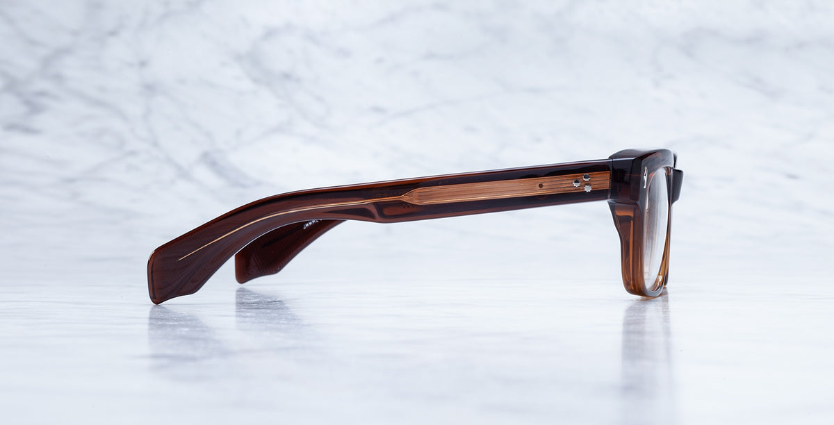 Side view of Jacques Marie Mage Molino in Hickory, featuring brown, thick-rimmed rectangular frames resting on white marble. Focus is on the arm and textured material of the eyeglasses.