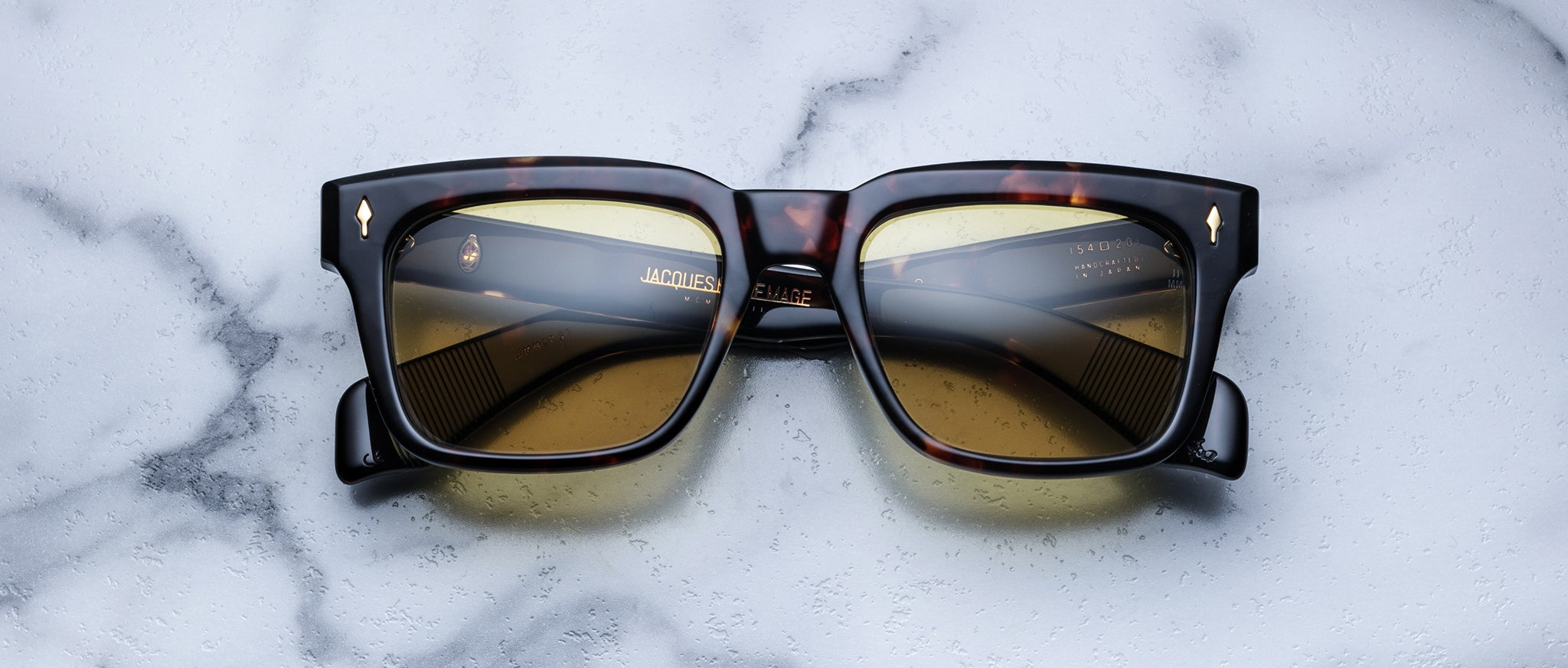The Jacques Marie Mage Torino, bold rectangular eyeglass frames in tortoiseshell with brown lenses by Jacques Marie Mage, rest on a white marble surface, evoking the essence of handcrafted acetate eyewear.