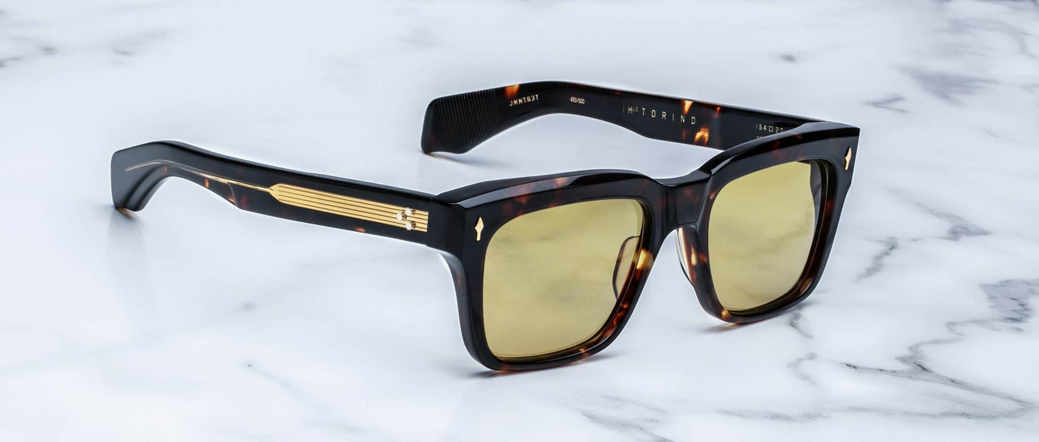A pair of Jacques Marie Mage Torino rectangular sunglasses with yellow lenses and gold arm detailing, handcrafted from acetate, rest on a white marble surface.