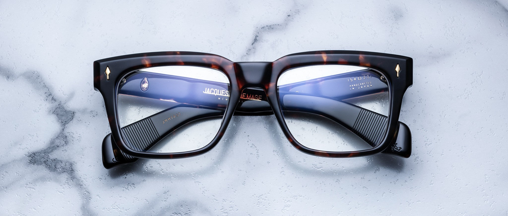 A pair of Jacques Marie Mage Torino bold rectangular eyeglass frames in tortoiseshell handcrafted acetate by Jacques Marie Mage rests on a white marble surface.