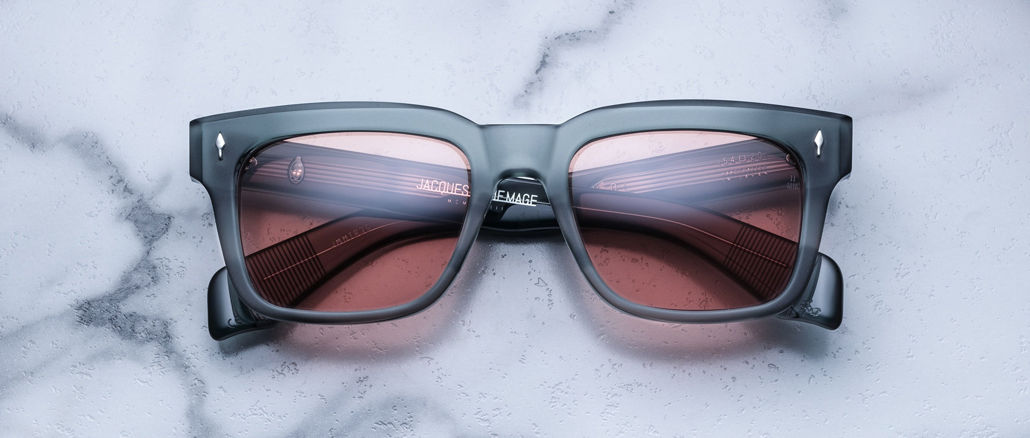 The Jacques Marie Mage Torino features bold rectangular gray acetate frames with pink-tinted lenses, shown resting on a white marble surface and reflecting the brand's signature handcrafted eyewear style.