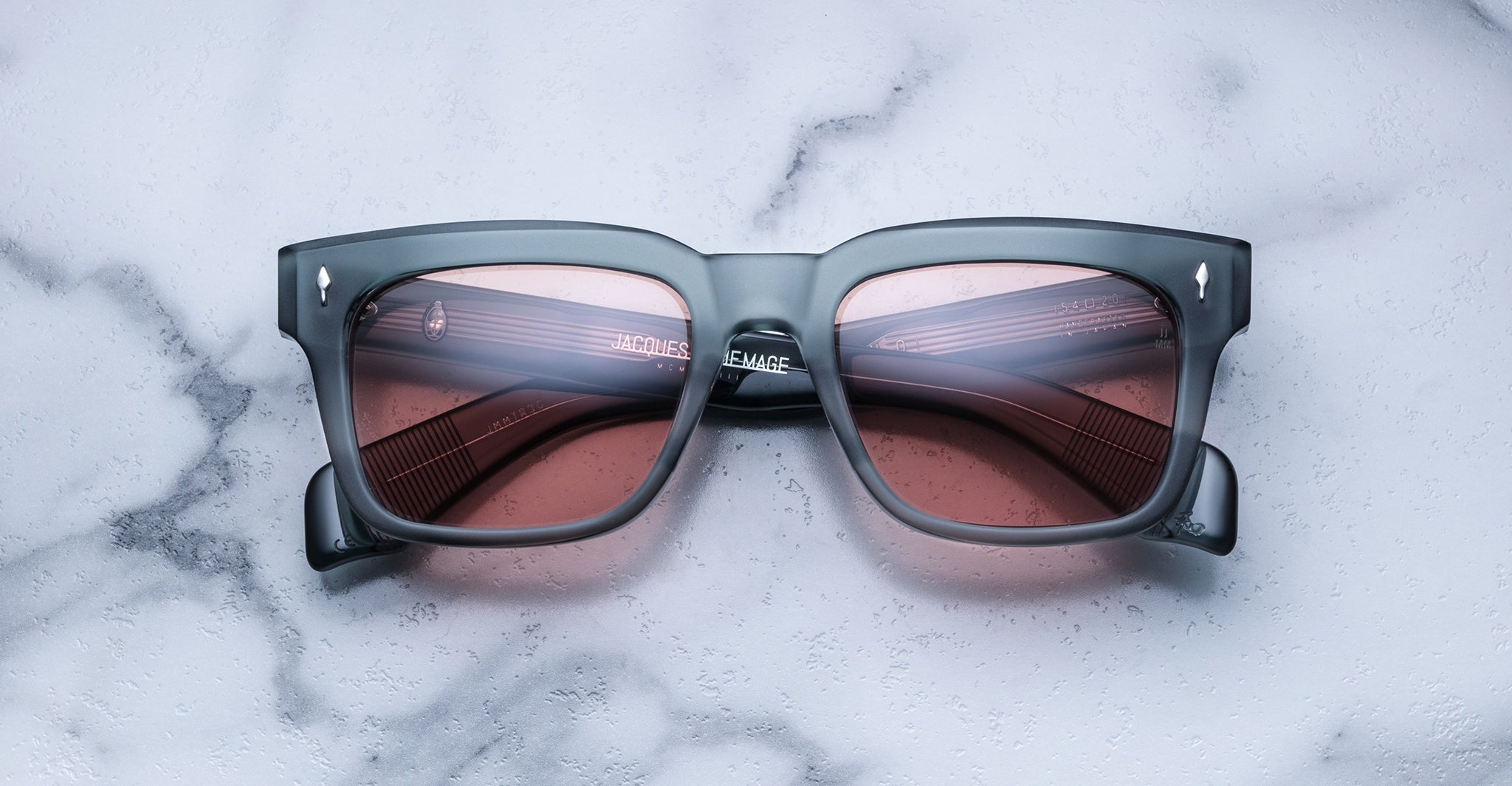 The Jacques Marie Mage Torino features bold rectangular gray acetate frames with pink-tinted lenses, shown resting on a white marble surface and reflecting the brand's signature handcrafted eyewear style.