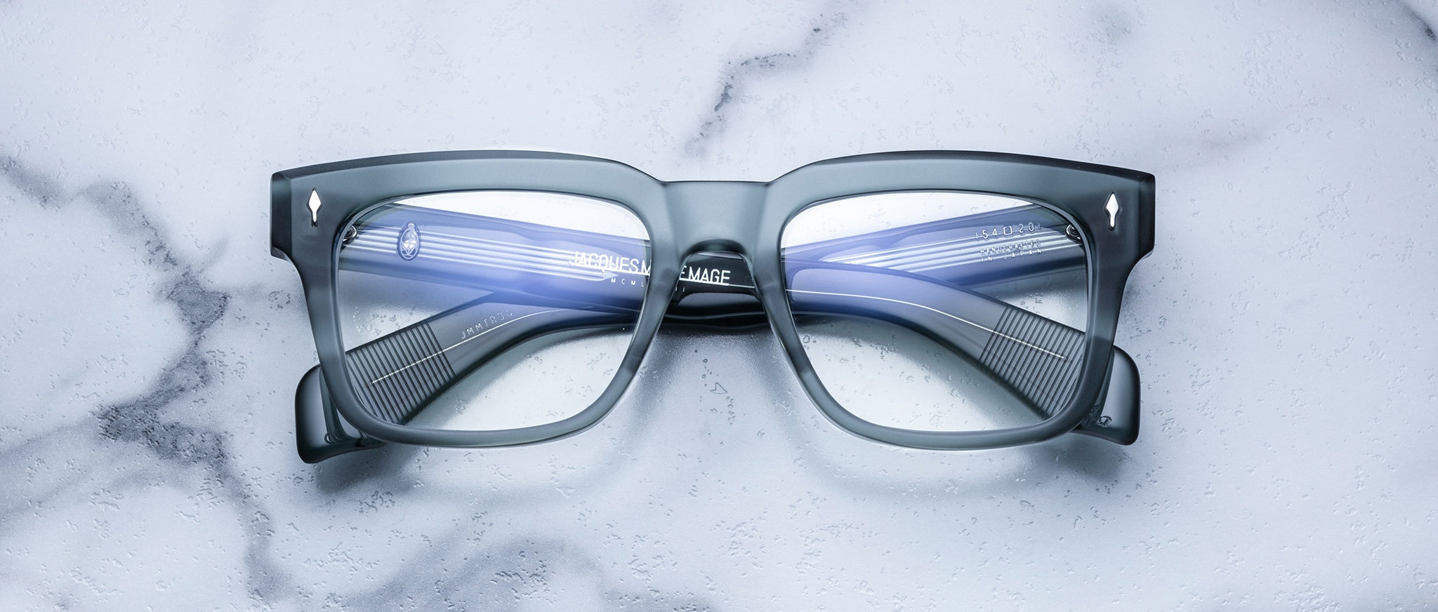 The Jacques Marie Mage Torino rectangular eyeglass frames, handcrafted from acetate by Jacques Marie Mage, rest on a white marble surface with reflections visible on their lenses.