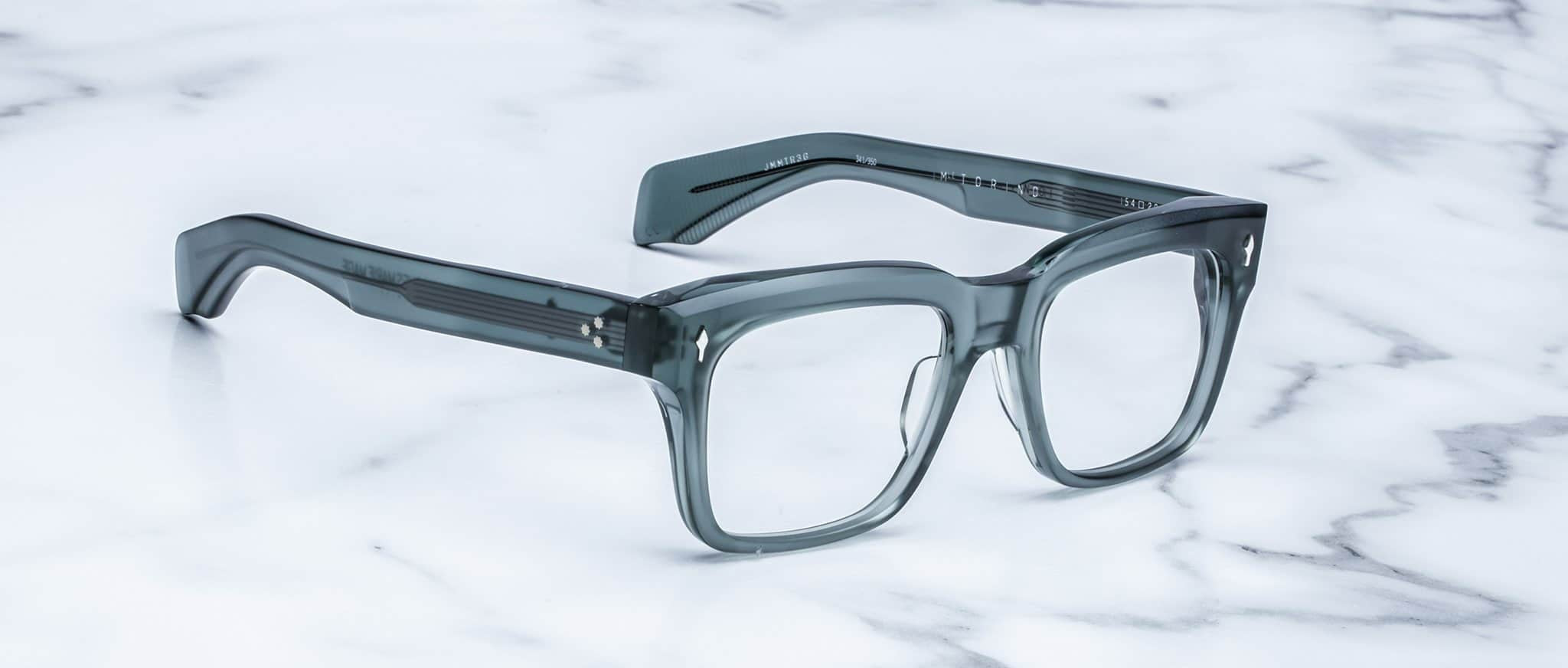 Jacques Marie Mage Torino eyewear: clear gray handcrafted acetate frames with bold rectangular design, inspired by the Jacques Marie Mage aesthetic, shown on a white marble surface.
