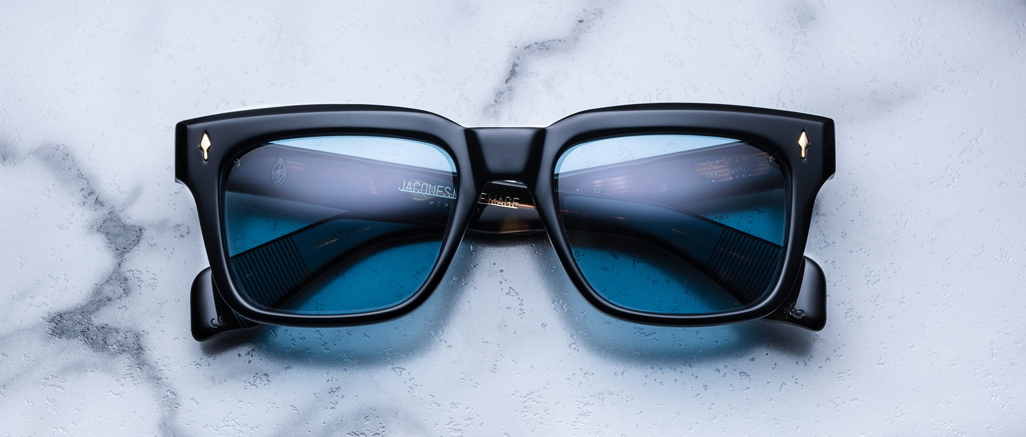 A pair of Jacques Marie Mage Torino sunglasses by Jacques Marie Mage, featuring black frames and blue-tinted lenses, rests on a white marble surface.