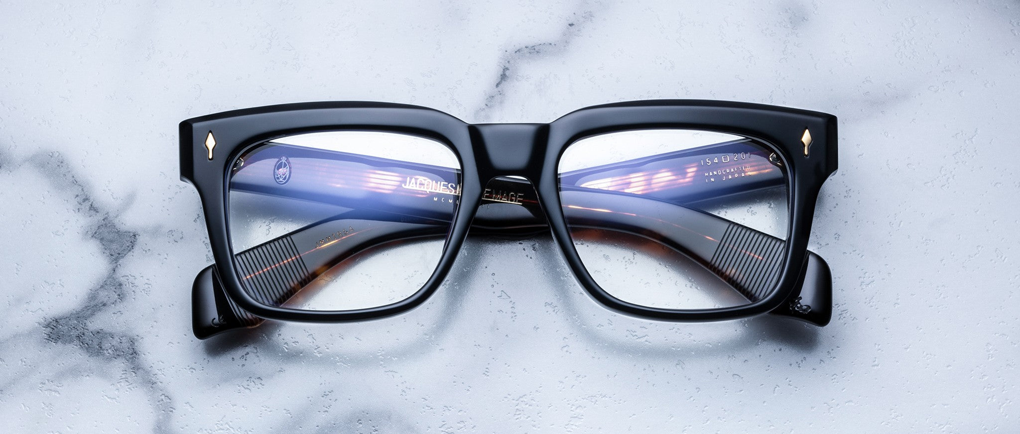 Jacques Marie Mage Torino eyeglasses feature black bold rectangular frames with gold corner accents, styled by Jacques Marie Mage, displayed on a white marble surface.