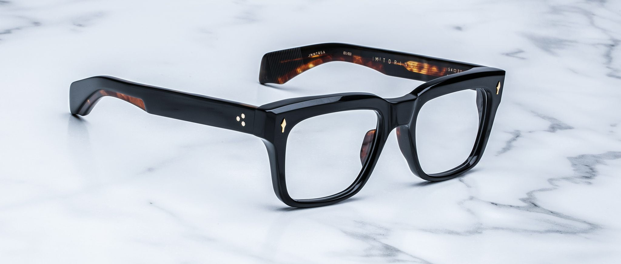 The Jacques Marie Mage Torino features bold rectangular acetate frames with thick black rims and subtle amber details, handcrafted by Jacques Marie Mage, displayed on a white marble surface.