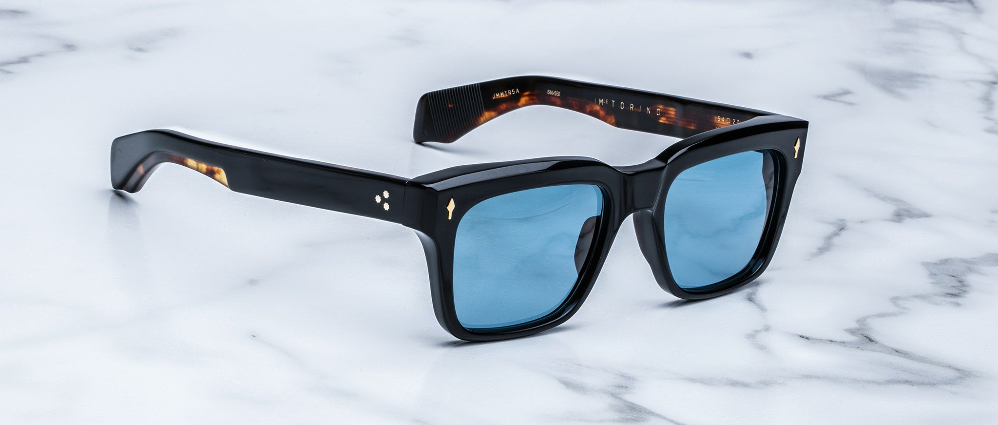 The Jacques Marie Mage Torino sunglasses by Jacques Marie Mage feature bold black square frames and blue tinted lenses, elegantly displayed on a white marble surface.